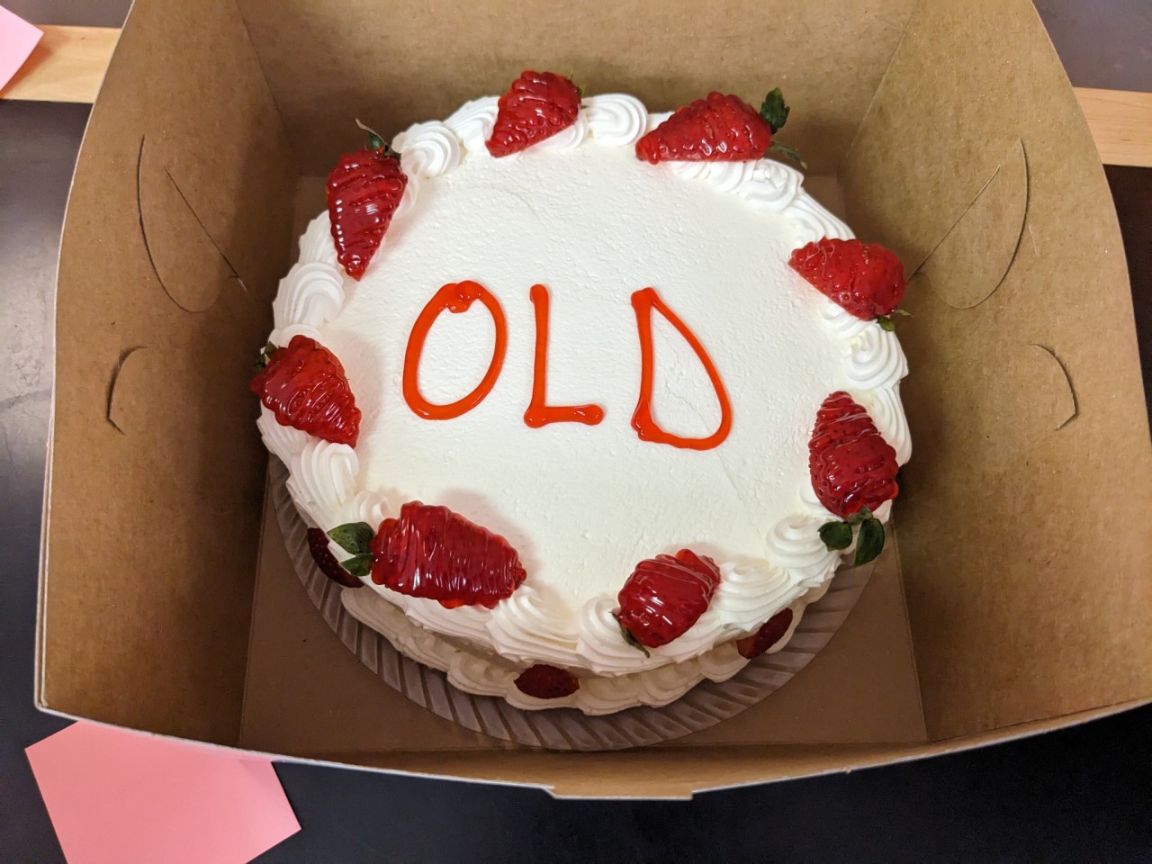 a lovely strawberry cake with white frosting sitting in a box, decorated with one word in all caps: OLD.